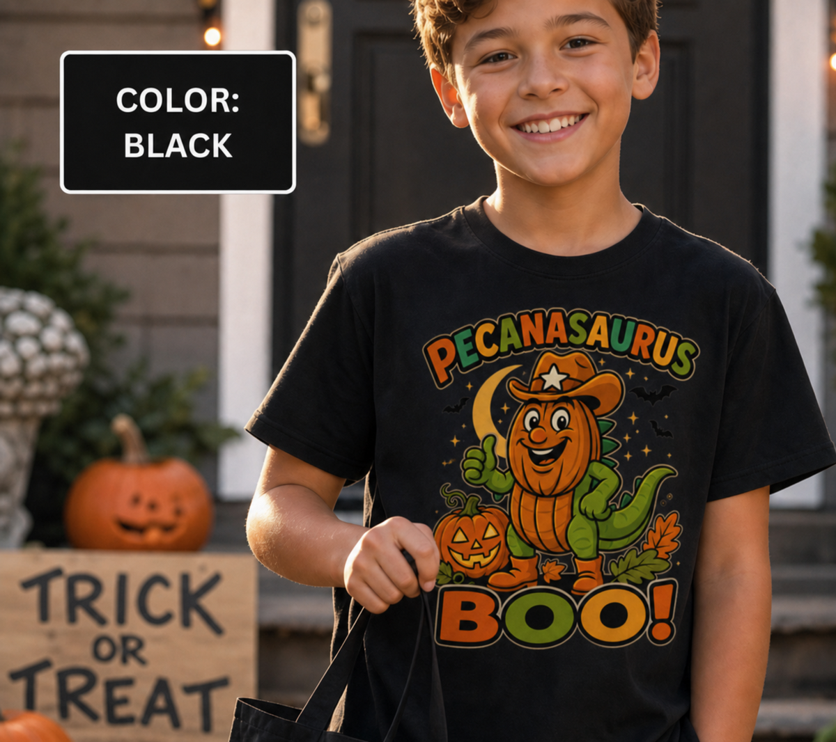 A smiling boy wearing a black graphic T-shirt with a colorful 'Pecanasaurus Boo!' Halloween design, standing in front of a decorated house. A black color label reading 'COLOR: BLACK' appears in the top left.