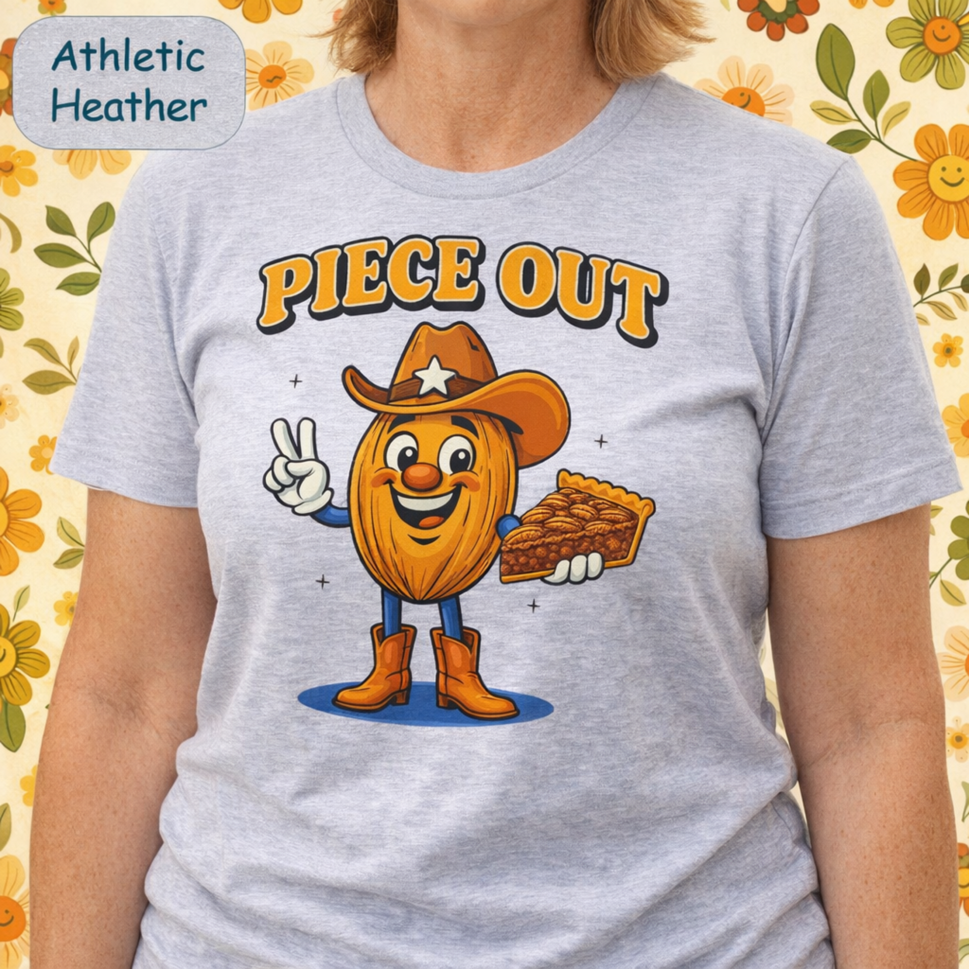 A person wearing a light heather gray graphic T-shirt with a cartoon pie character dressed as a cowboy and the words “PIECE OUT” above it. A badge in the top left reads “Athletic Heather.