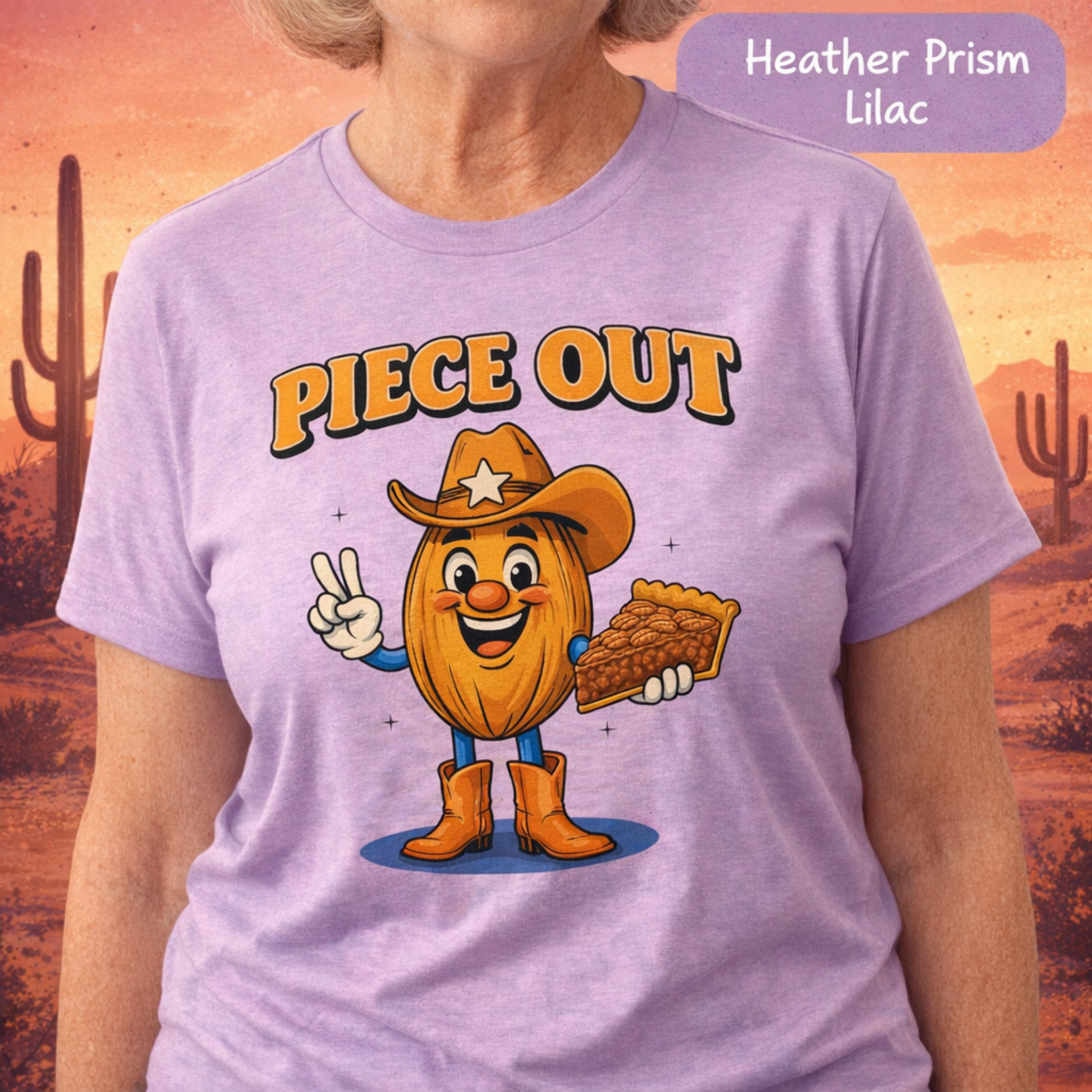 A person wearing a light lavender heather T-shirt with a cartoon graphic of an acorn character dressed as a cowboy holding a pie slice, with the text 'PIECE OUT' above. A purple label in the top right reads 'Heather Prism Lilac'.
