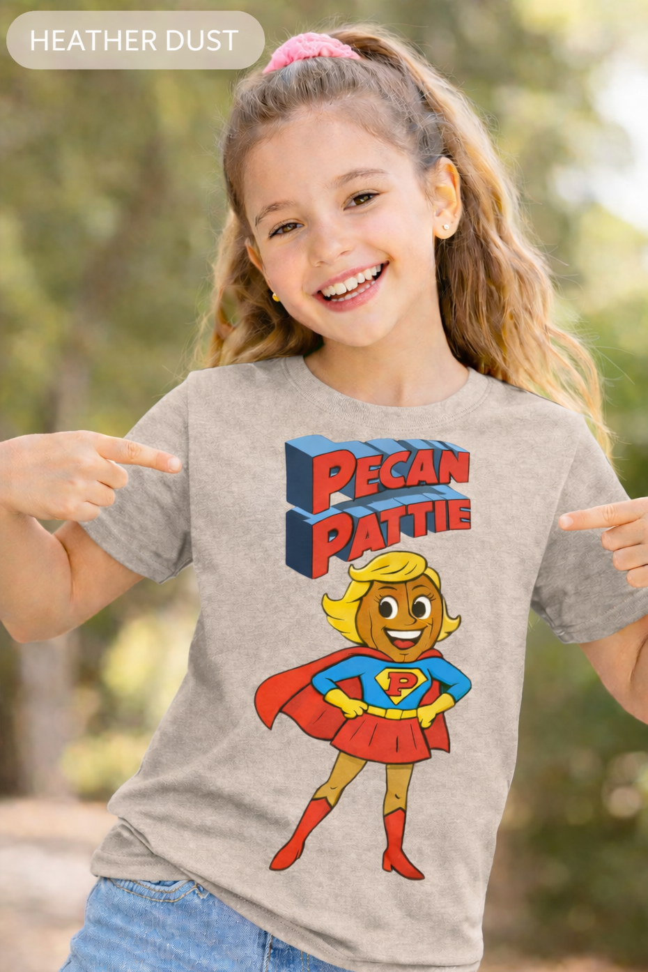 Happy girl modeling a Bella+Canva youth T-shirt featuring the Super Pecan Pattie cartoon superhero design, pointing at the graphic with an outdoor background.