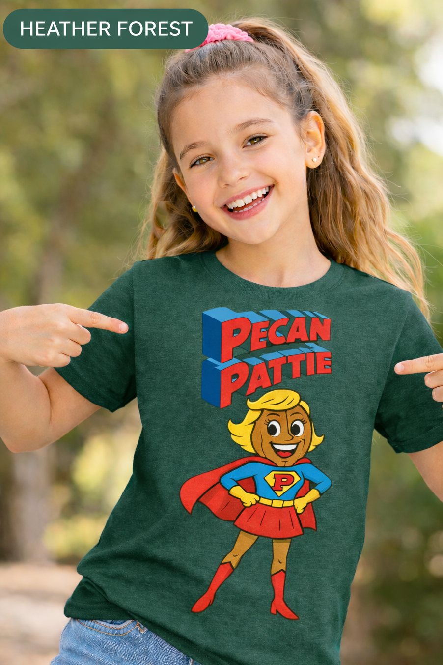 Happy girl modeling a Bella+Canva youth T-shirt featuring the Super Pecan Pattie cartoon superhero design, pointing at the graphic with an outdoor background.