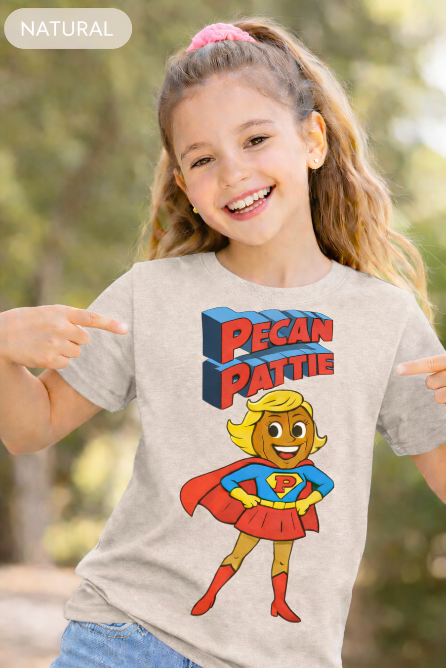 Happy girl modeling a Bella+Canva youth T-shirt featuring the Super Pecan Pattie cartoon superhero design, pointing at the graphic with an outdoor background.