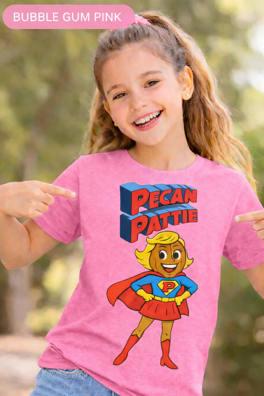 Happy girl modeling a Bella+Canva youth T-shirt featuring the Super Pecan Pattie cartoon superhero design, pointing at the graphic with an outdoor background.