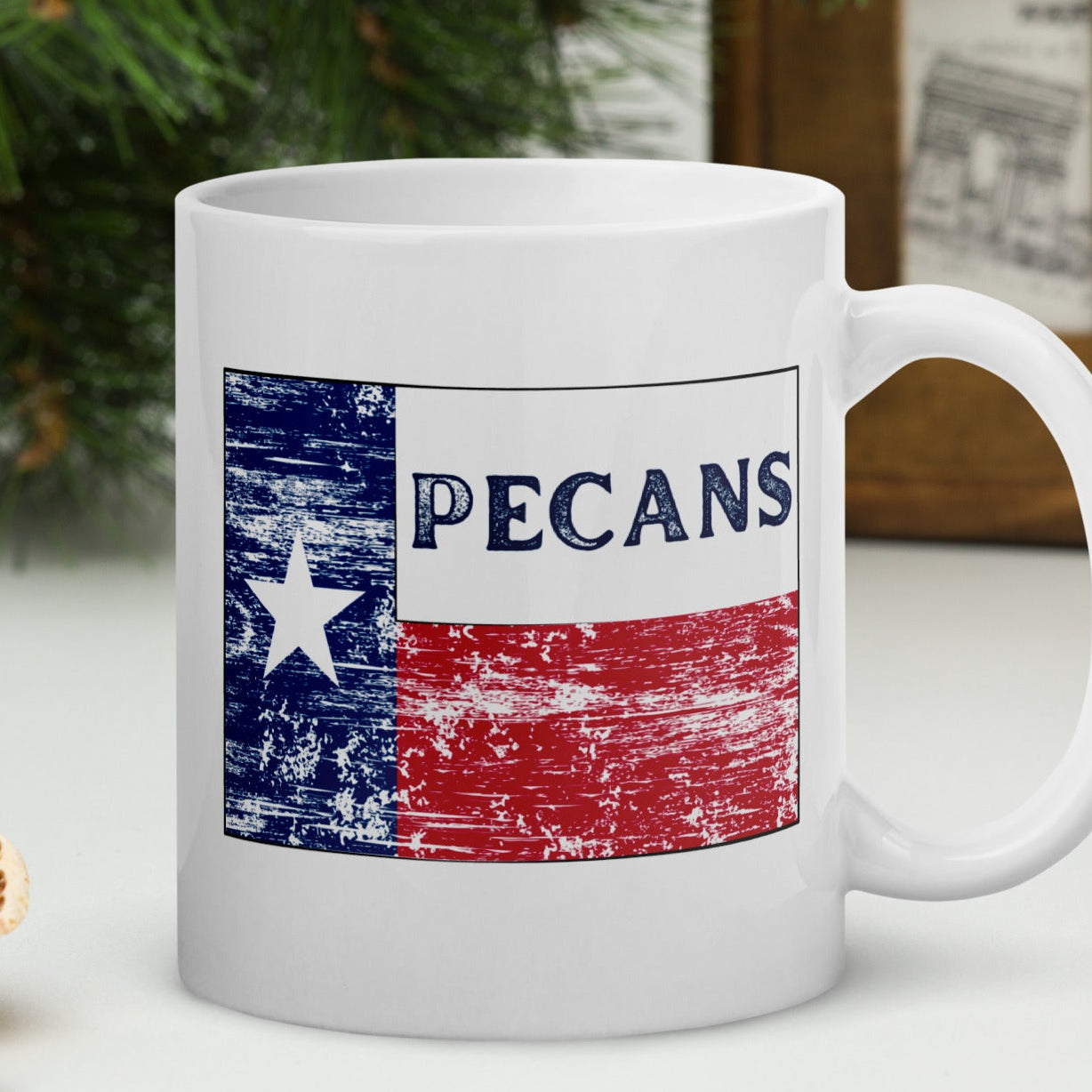 White ceramic mug with a distressed Texas flag design and the word “PECANS,” sitting on rustic wooden table with Texas-themed background.